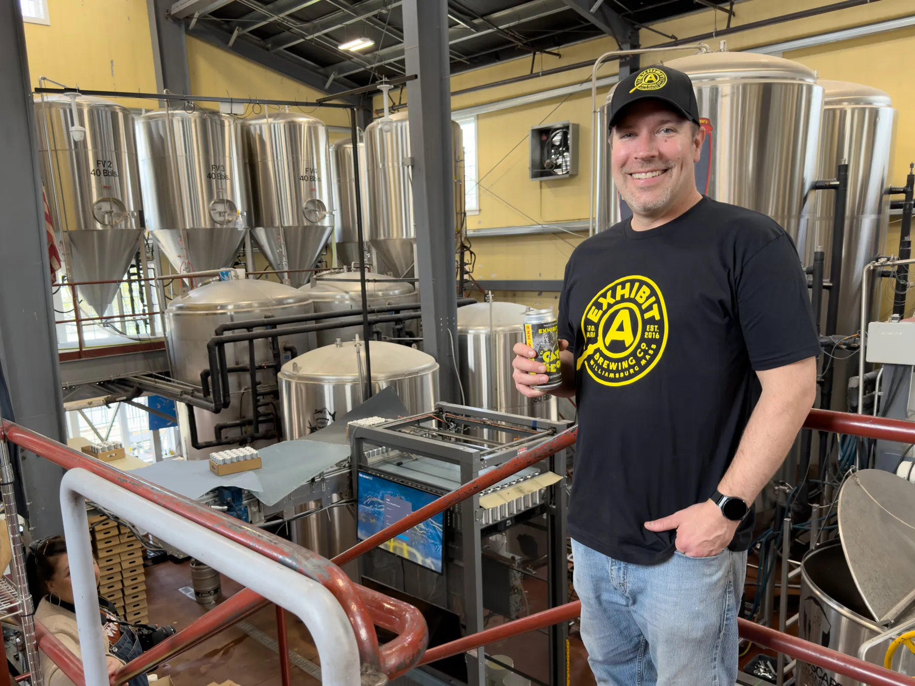 Exhibit A's brewmaster, Dennis holding a beer can in front of the brewery's fermentation tanks
