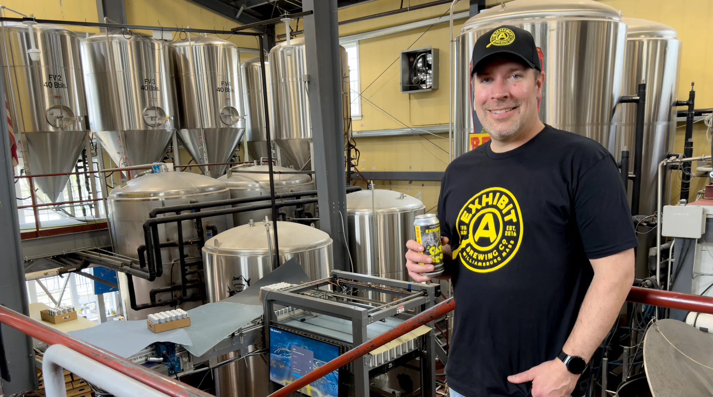 Exhibit A's brewmaster, Dennis holding a beer can in front of the brewery's fermentation tanks